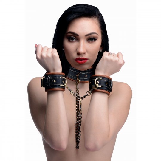 A luxurious vegan-friendly collar and wrist restraint set featuring a brown PU leather collar with three D-rings, two matching cuffs with D-rings, and chain connectors, all accented with brass toned hooks and gold embellishments.