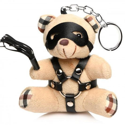 A cute beige bondage bear with light peach fur, wearing a faux leather harness with metal O-rings, holding a mini flogger, and featuring a removable eye mask, designed to be a keychain accessory or novelty gift.